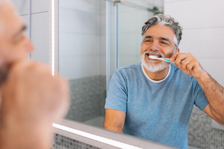 A gray-haired man in his 50s or 60s stands before a bathroom mirror while he brushes his teeth