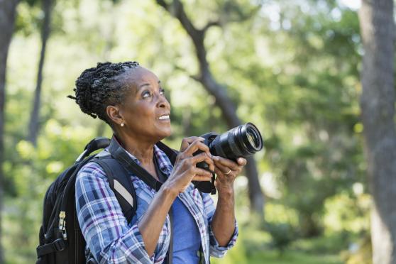 A woman in a blue plaid shirt holds a DSLR camera toward a subject in the woods. She's wearing a backpack and photographing nature.