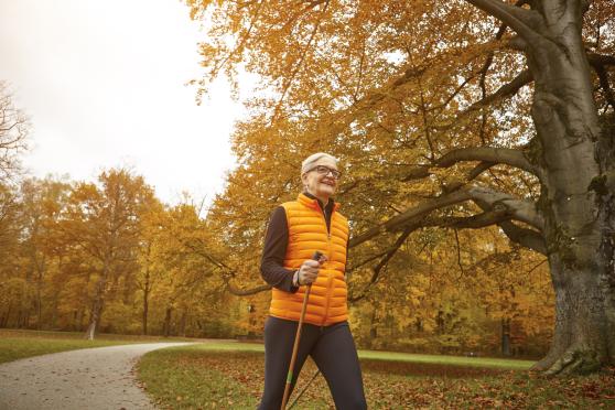 An older woman smiles as she strolls along a path on a beautiful autumn day, with lots of yellow foliage in the background. She's wearing a bright orange puffy vest and walking with a wooden cane.