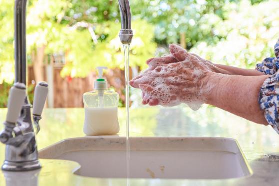 A woman's hands and forearms are visible as she washes her hands with soap and water at a white sink.