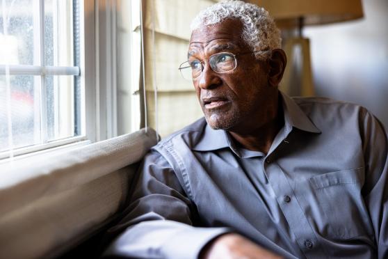 An older man looks out the window with a melancholy expression on his face.