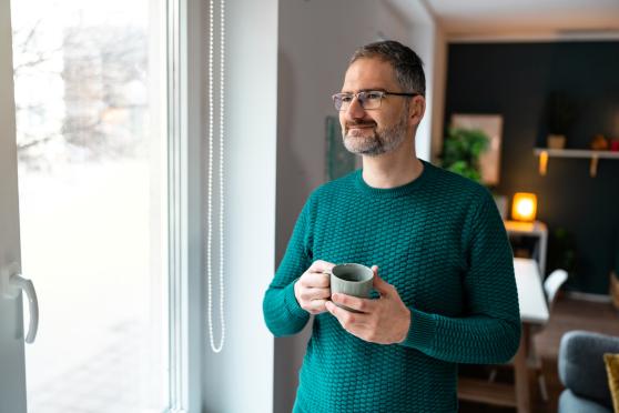 A middle-aged man stands in his home holding a coffee mug, looking out the window.