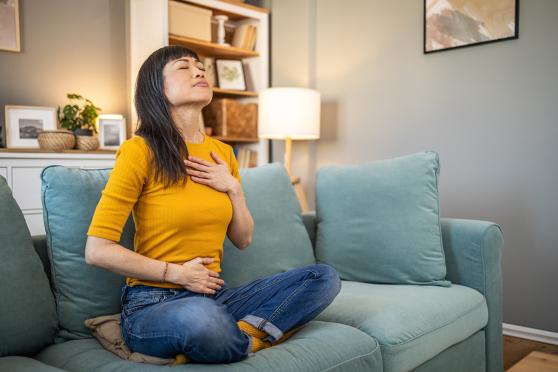 A woman with long, dark hair and bangs is meditating while sitting crosslegged on a blue sofa with one hand on her chest and the other on her belly.