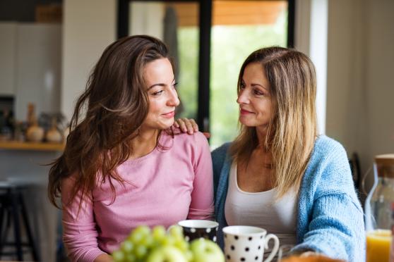 Two women sit side by side at a kitchen table, two mugs in front of them. The woman in the blue shirt rests her hand supportively on the shoulder of the woman in the pink shirt. They are both smiling slightly.