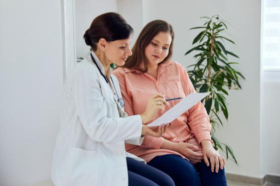 A woman sits on an exam table beside a doctor, who is pointing at a piece of paper and explaining something to her.